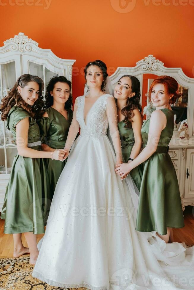 Enjoying the preparations for the wedding. bridesmaids helping the bride to put on the veil and dress and smiling while standing in the room photo