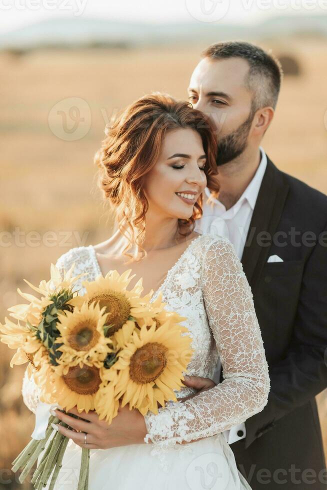 Wedding portrait of the bride and groom. The groom hugs the bride from behind, next to a bale of hay. Red-haired bride in a long dress with a bouquet of sunflowers. Stylish groom. Summer photo