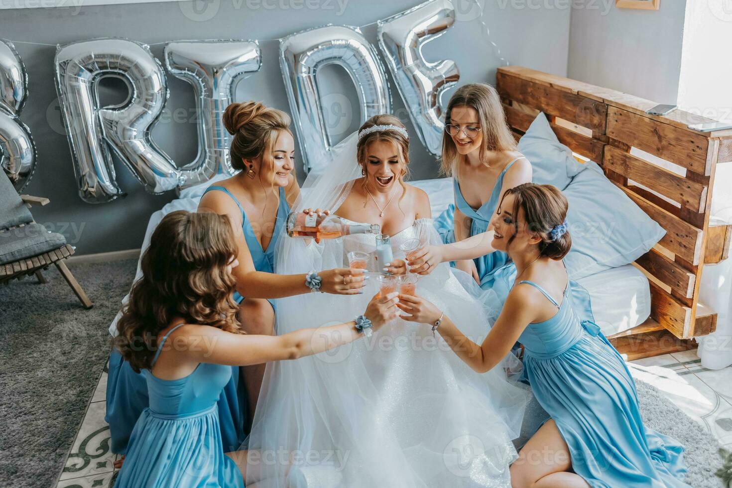 joven damas de honor en azul seda vestidos bebida champán en el de la novia habitación. hermosa mujer celebrando despedida de soltera fiesta sentado en cama y con champán. foto