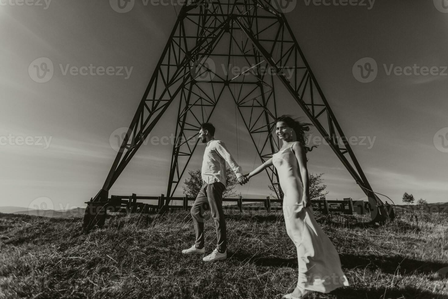 Stylish model couple in the mountains in summer. A young boy and girl in a white silk dress are standing near large structures of power lines. Black and white contrast photo