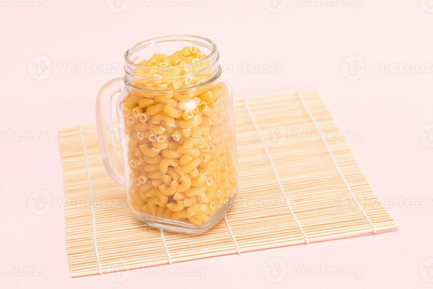 Uncooked Chifferi Rigati Pasta in Glass Jar on Bamboo Mat on Pink Background. Fat and Unhealthy Food. Scattered Classic Dry Macaroni. Italian Culture and Cuisine. Raw Pasta photo