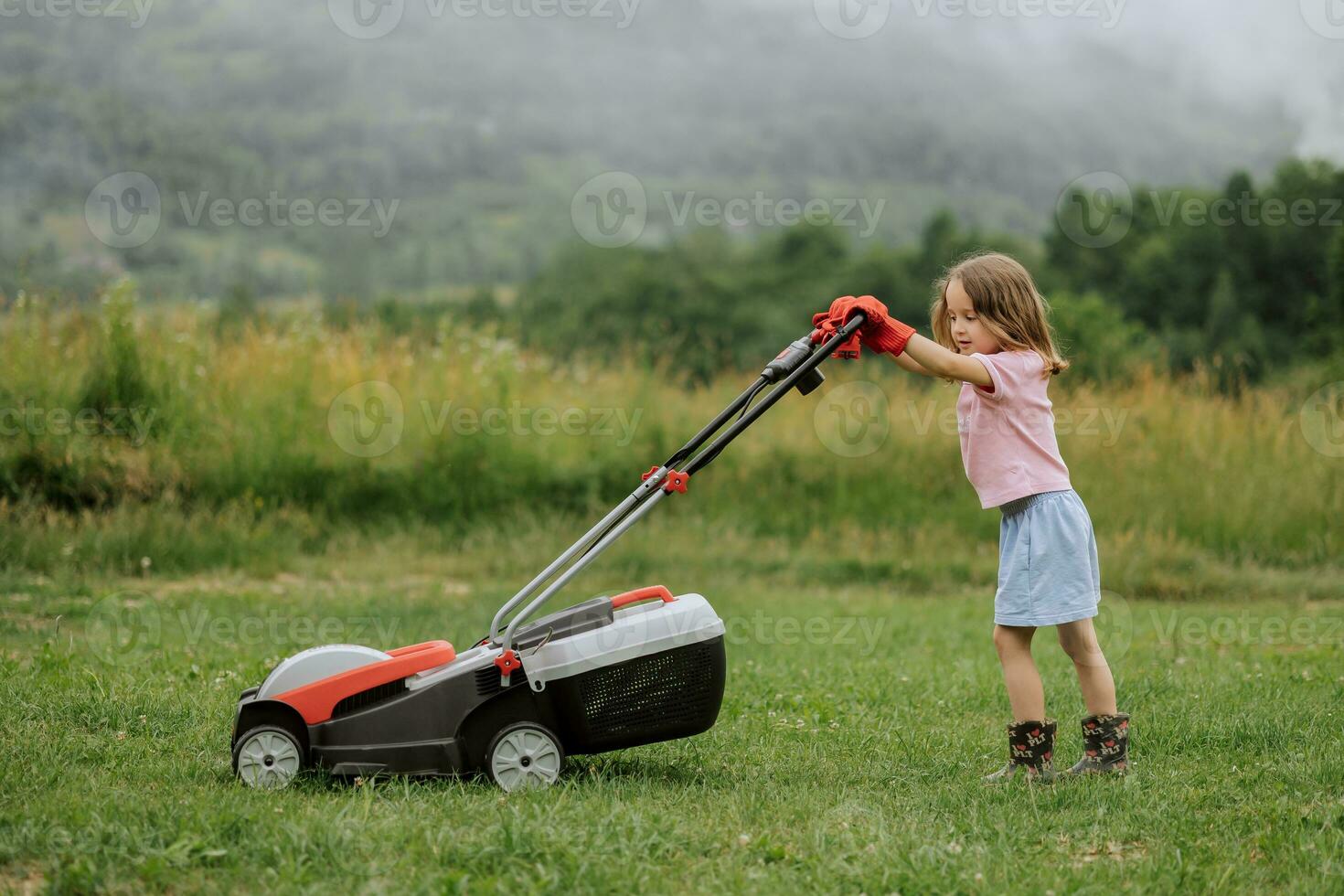 A child in boots in the form of a game mows grass with a lawnmower in the yard against the background of mountains and fog, the concept of garden tools photo