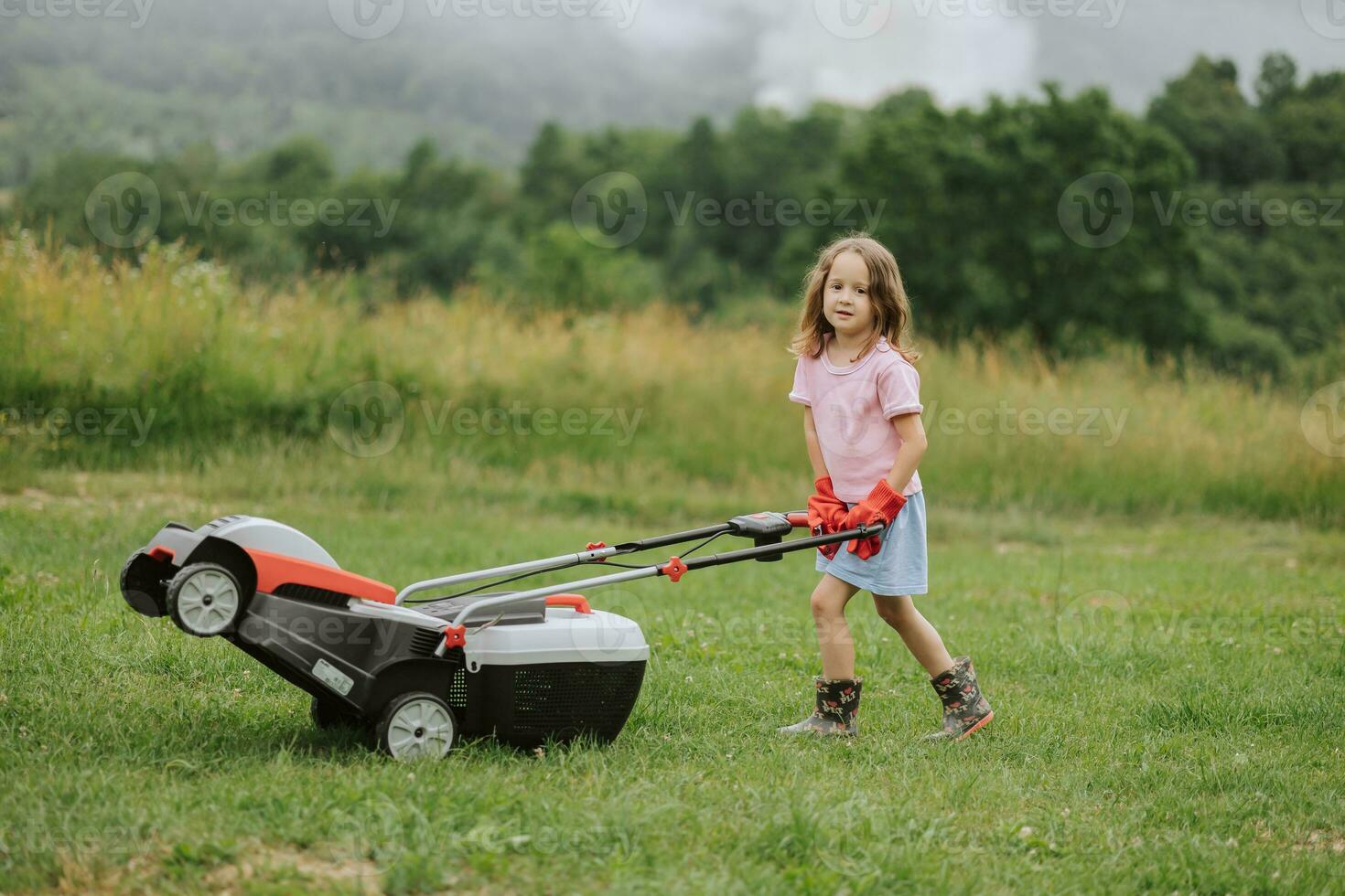 A child in boots in the form of a game mows grass with a lawnmower in the yard against the background of mountains and fog, the concept of garden tools photo