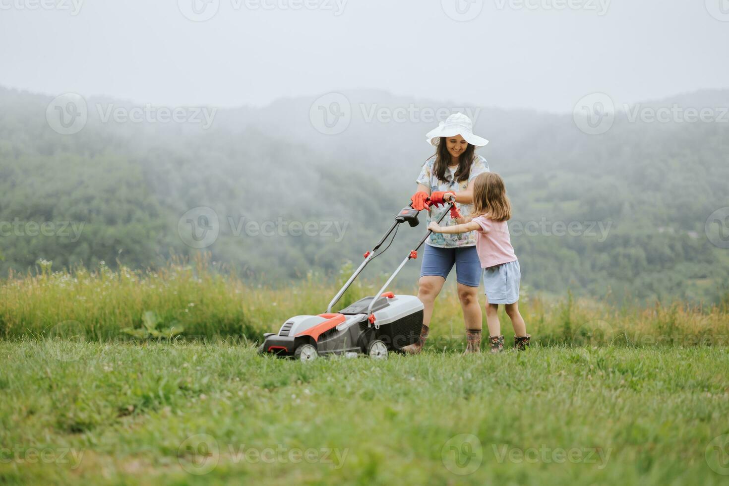 A woman in boots with her child in the form of a game mows the grass with a lawnmower in the garden against the background of mountains and fog, garden tools concept, work, nature. photo