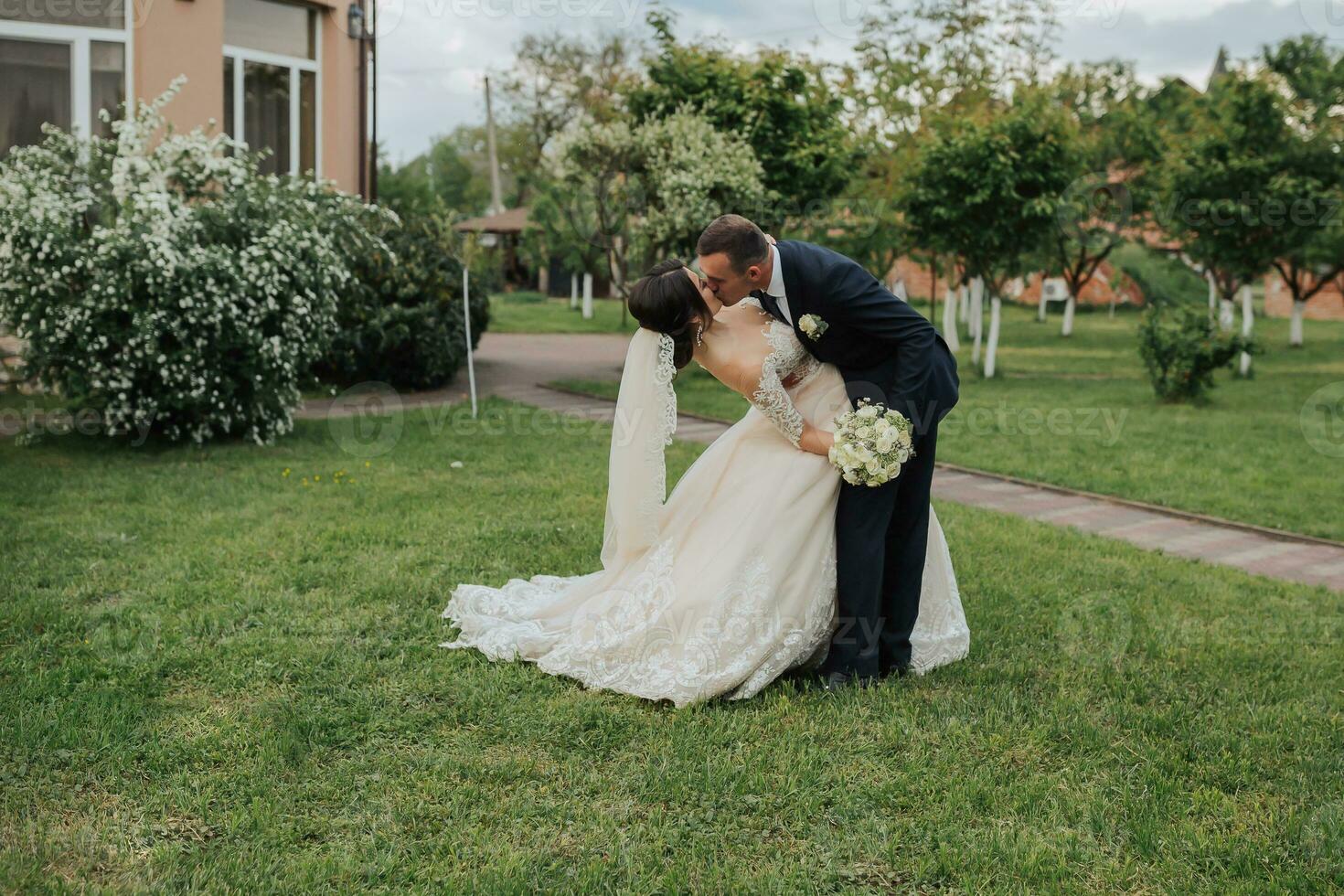 Portrait of the bride and groom in nature. Stylish bride and groom in a long lace dress are hugging and kissing near the trees in the garden. A happy couple in love photo