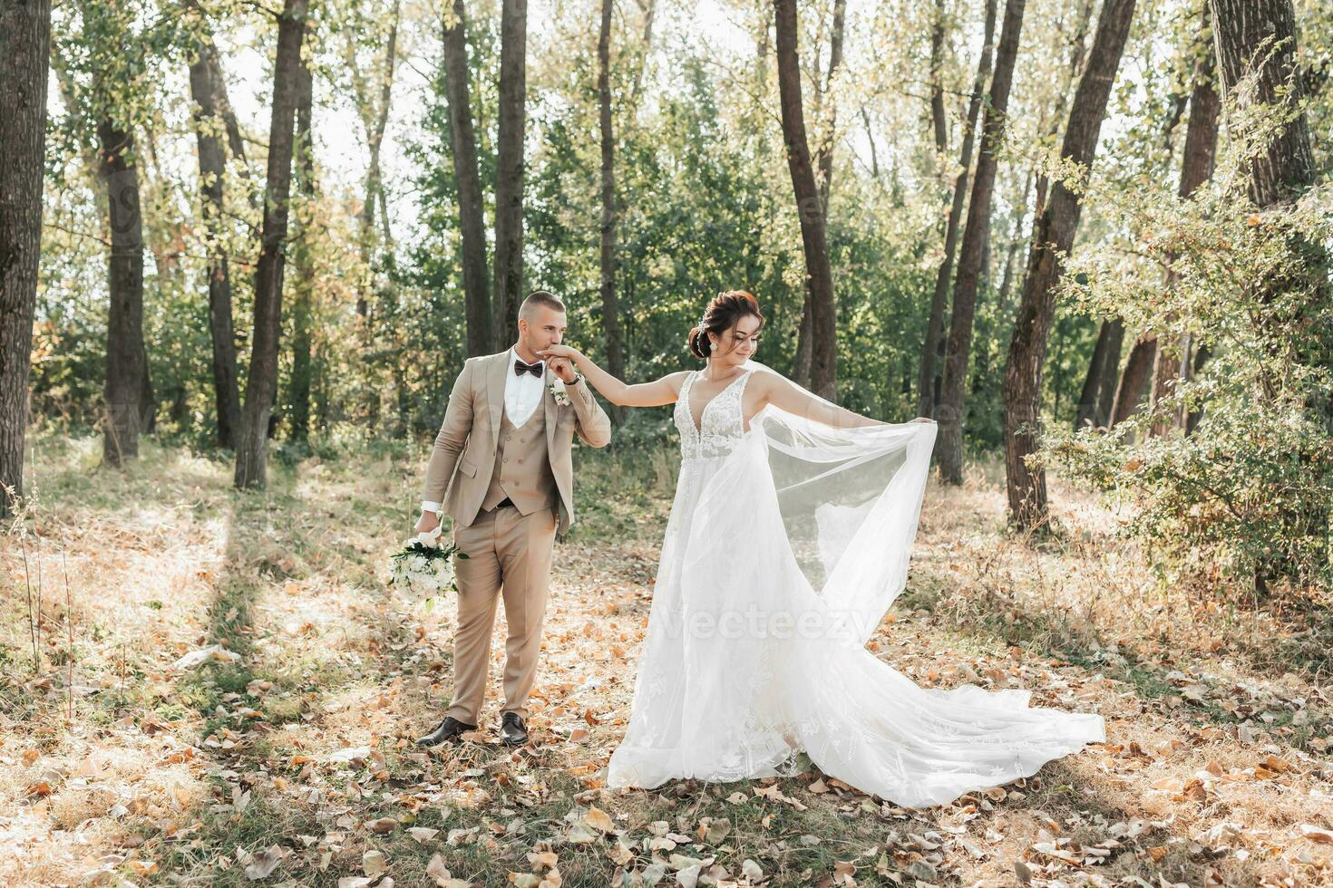 Wedding photo. The bride and groom are walking in the forest. The groom holds a bouquet and kisses the hand of his beloved, and she beautifully throws up her long veil. Couple in love. Summer light. photo