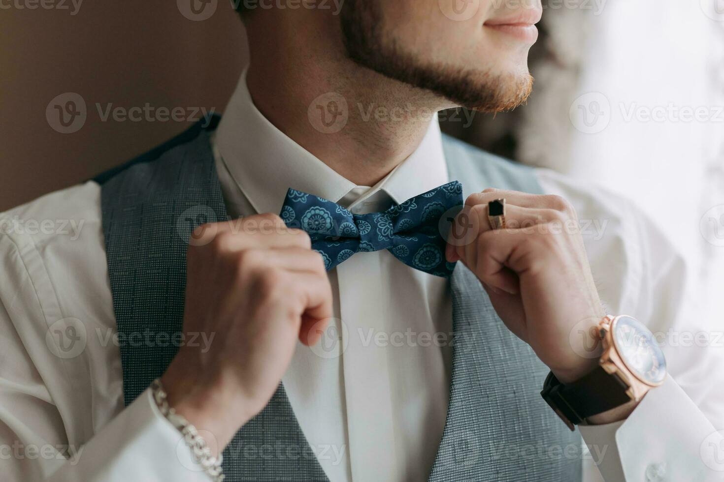 A man in a white shirt and a gray vest poses by the window and looks to the side, adjusts his blue bow tie. Front view. A stylish watch. Men's style. Fashion. Business photo