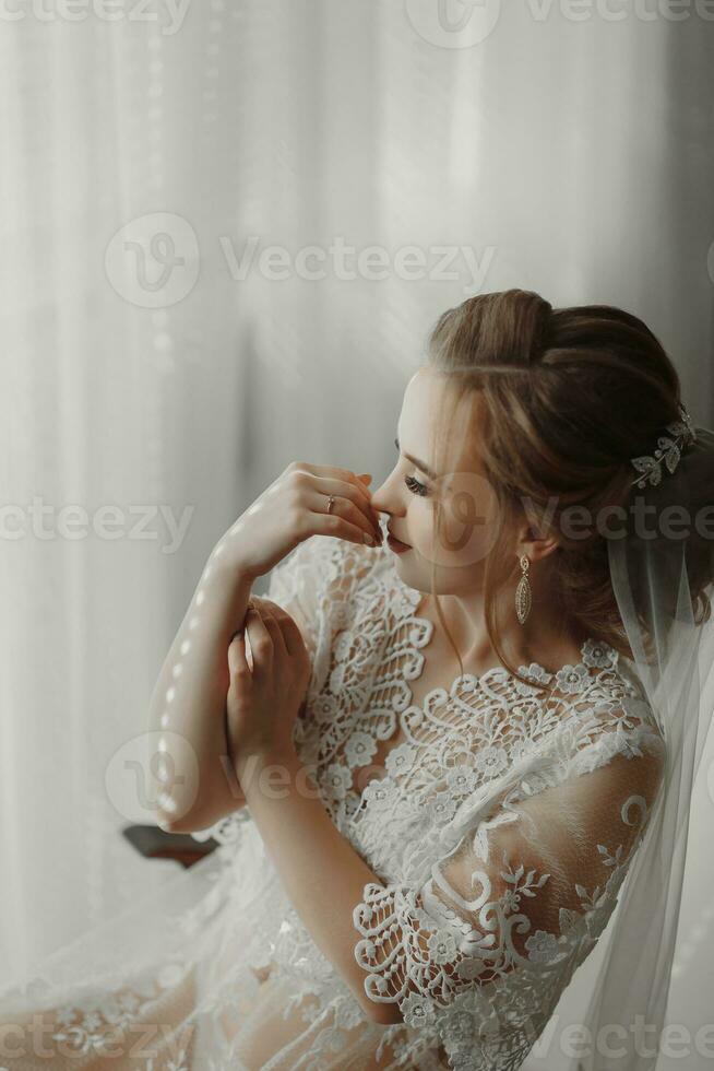 the bride in a transparent robe sits on the edge of a soft pink chair in a bright room, wears an elegant gold earring photo