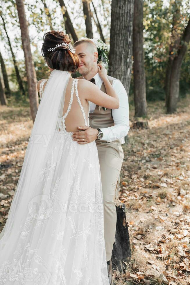 Wedding photo in nature. The bride hugs and kisses the groom. The bride has her shoulders turned. Beautiful hairstyle and hair decoration. Portrait of the bride and groom in the forest