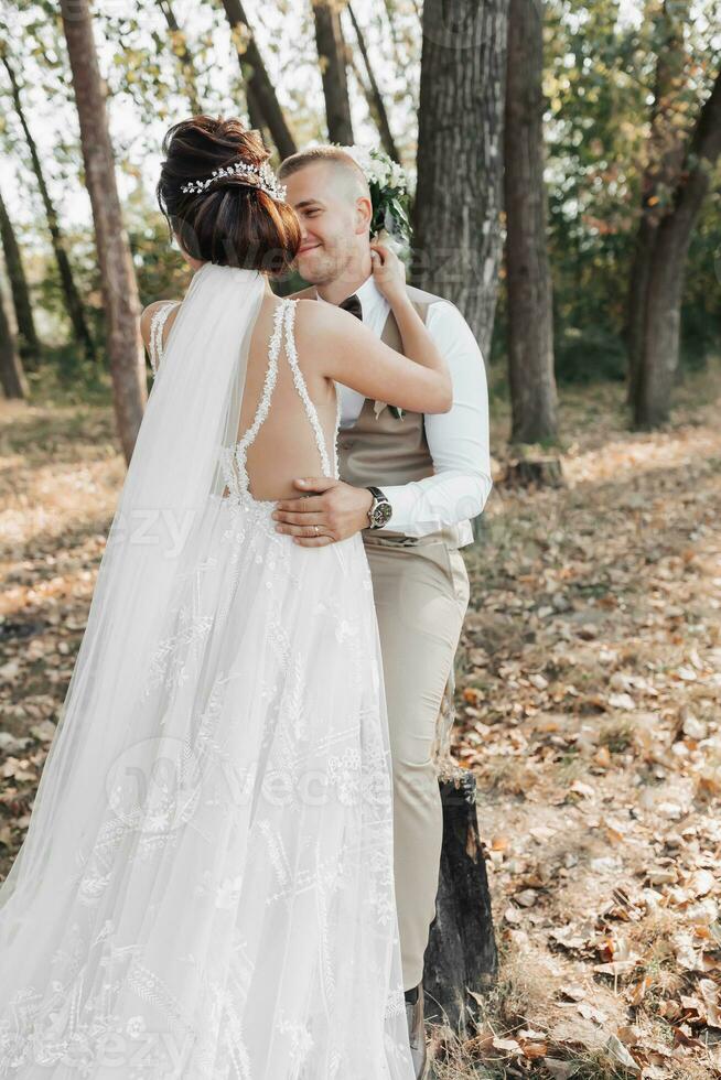 Boda foto en naturaleza. el novia abrazos y Besos el novio. el novia tiene su espalda transformado. hermosa peinado y pelo decoración. retrato de el novia y novio en el bosque