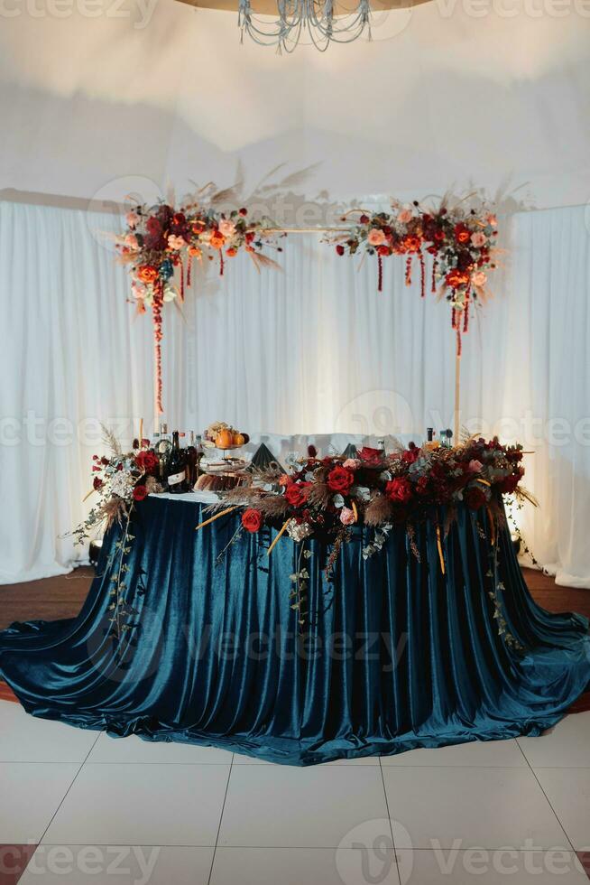 The wedding table of the bride and groom, decorated with red flowers, blue velor, is made in red and blue. Flowers on stands. Wedding details. light photo