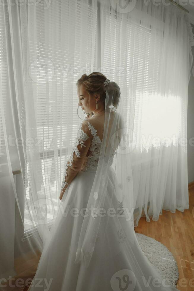 Preparation for the wedding. Beautiful young bride in white wedding dress indoors. Luxury model looks at wedding shoes, at home in studio room with big window. The girl shows a photo from the back