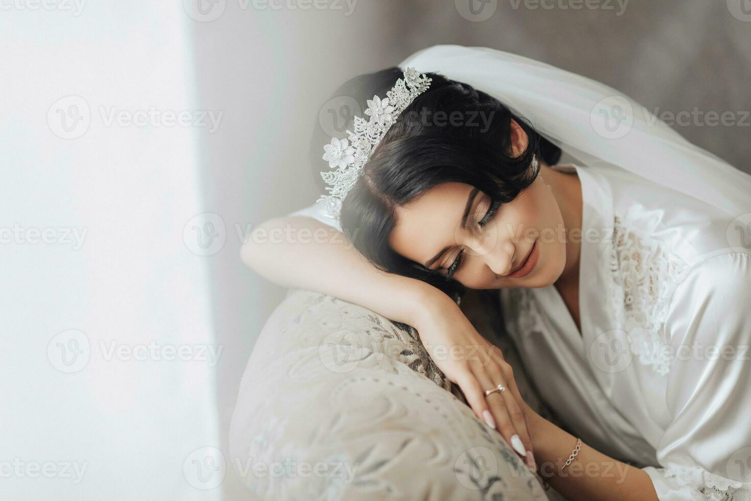 Portrait. A brunette bride in a robe and a chic crown is lying on the back of the sofa with her eyes closed. Gorgeous make-up and hair. Voluminous veil. Wedding photo. Beautiful bride photo