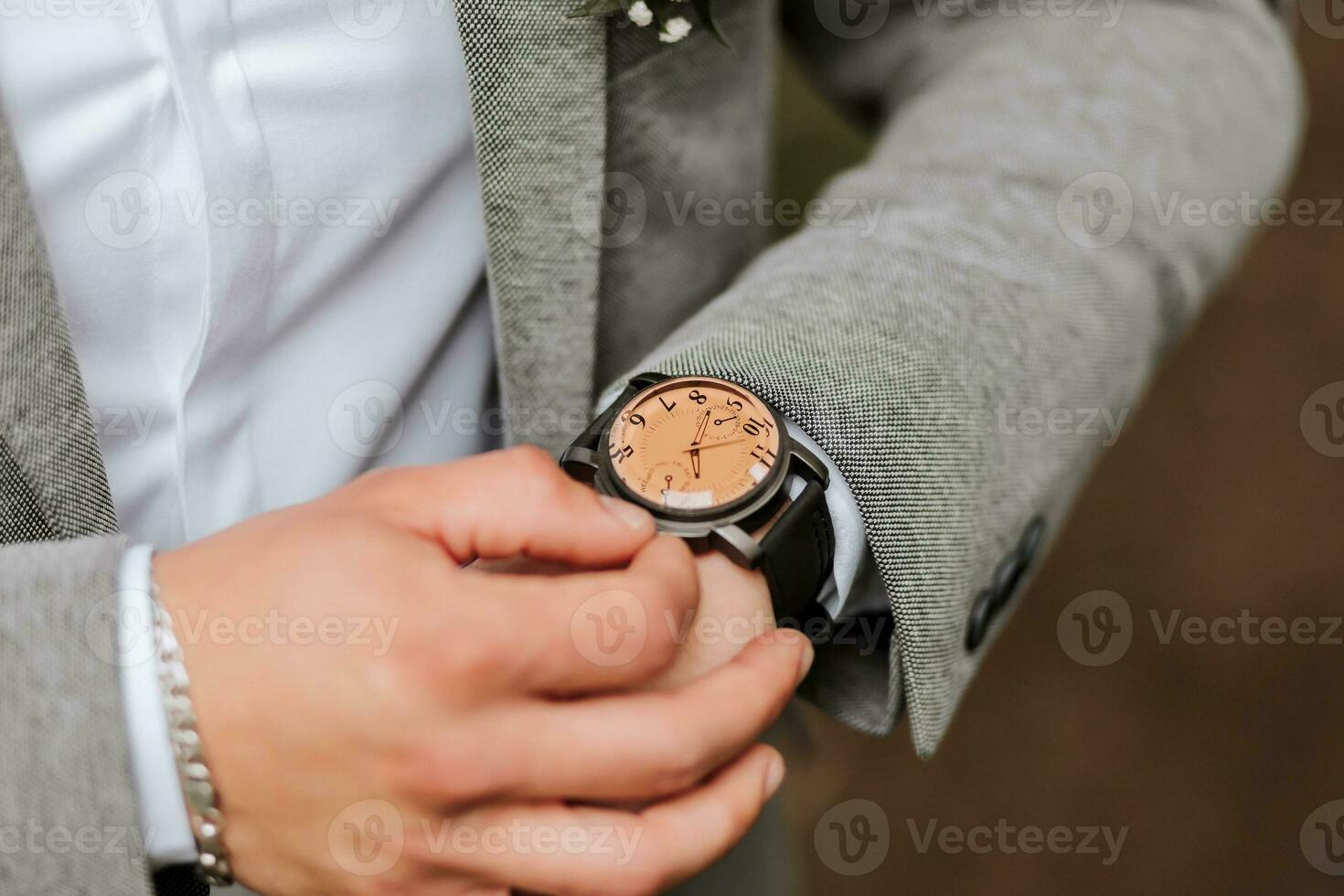 the stylish groom adjusted his watch. Luxury expensive watch on hand. Gray jacket, white shirt, ankle boots made of fresh flowers. Photo details