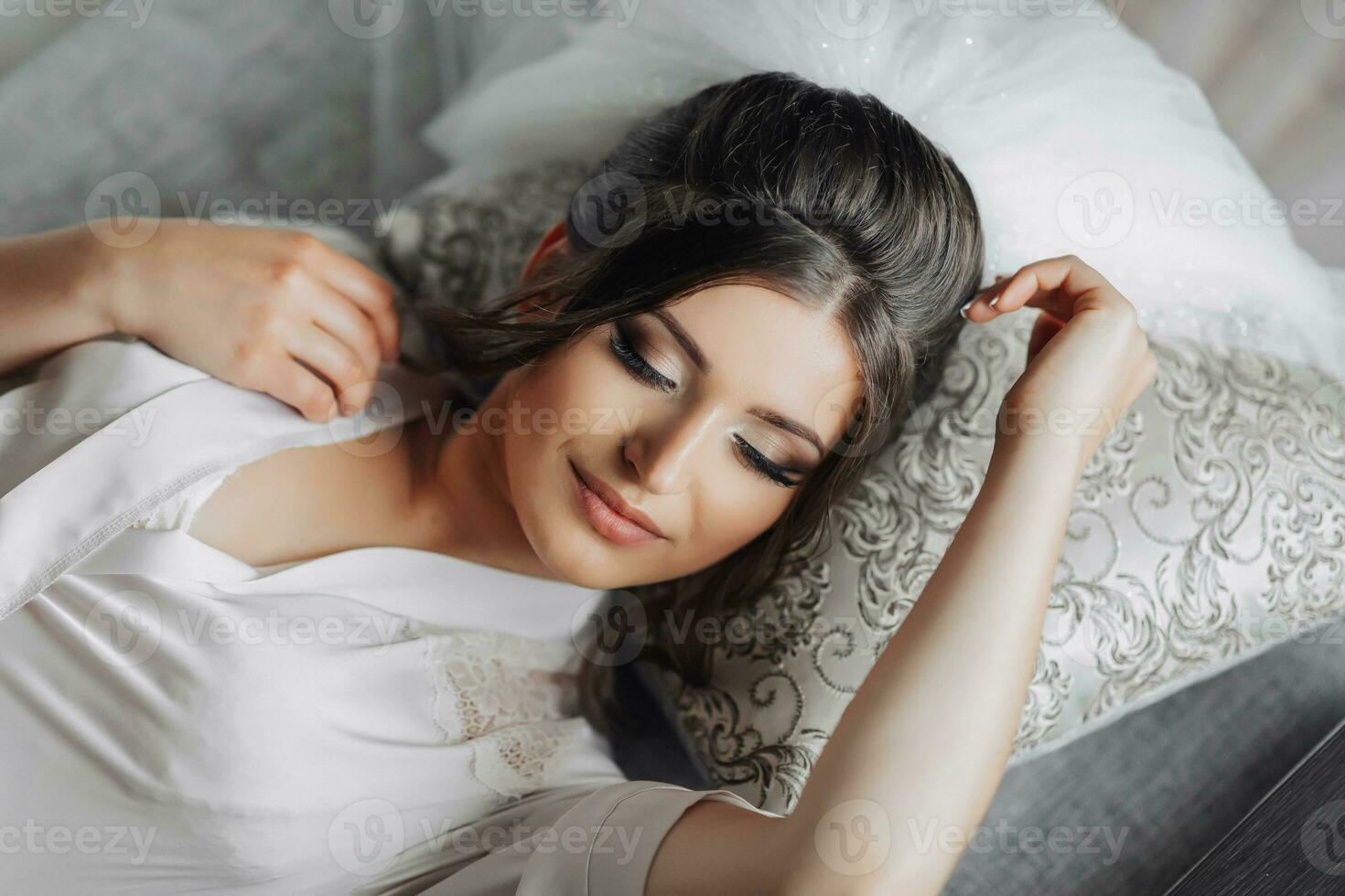 Brunette bride in a dressing gown posing for the camera while lying on a gray sofa. Portrait photo. Elegant hairstyle. Nice makeup. Beautiful hands. Morning of the bride. photo