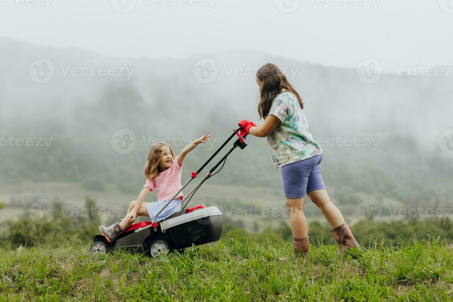 A woman in boots with her child in the form of a game mows the grass with a lawnmower in the garden against the background of mountains and fog, garden tools concept photo
