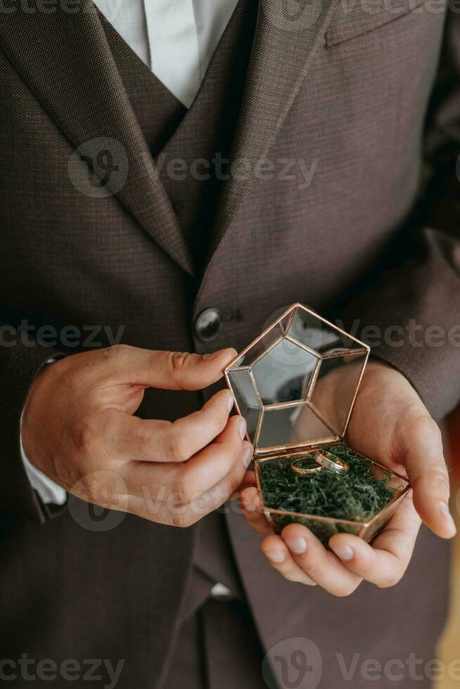 Wedding gold rings on a decorative glass box with wooden green moss, standing in the hands of the groom, close-up. Jewelry concept photo