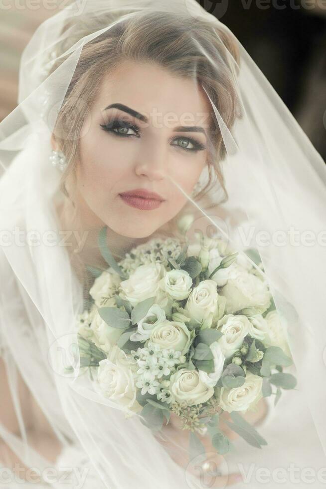 Curly blonde bride in a white dress, covered with a veil, poses for the camera with a bouquet of roses. Portrait of the bride. Beautiful makeup and hair. Wedding in nature photo