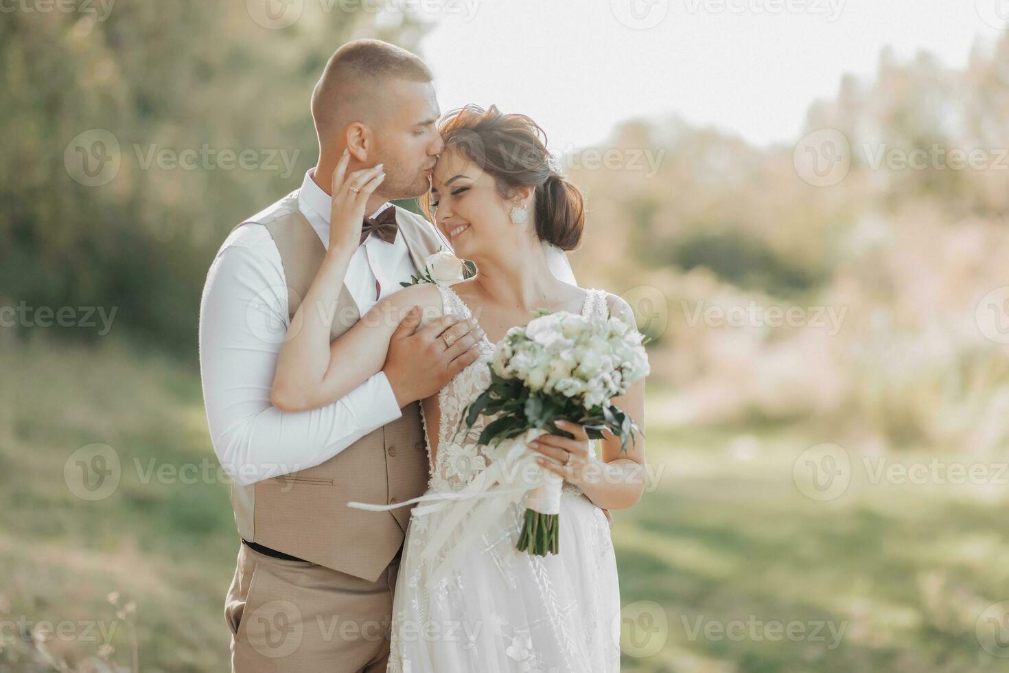 Wedding photo in nature. The bride and groom are standing in the forest, the groom hugs his beloved from behind and kisses her, she smiles sincerely. Portrait. Summer wedding