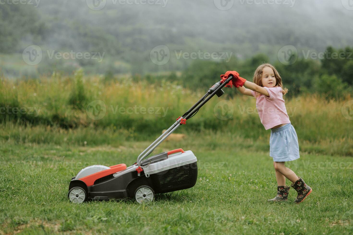 A child in boots in the form of a game mows grass with a lawnmower in the yard against the background of mountains and fog, the concept of garden tools photo