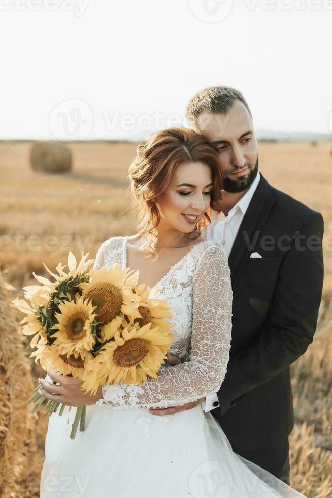 Wedding portrait of the bride and groom. The groom hugs the bride from behind, next to a bale of hay. Red-haired bride in a long dress with a bouquet of sunflowers. Stylish groom. Summer photo