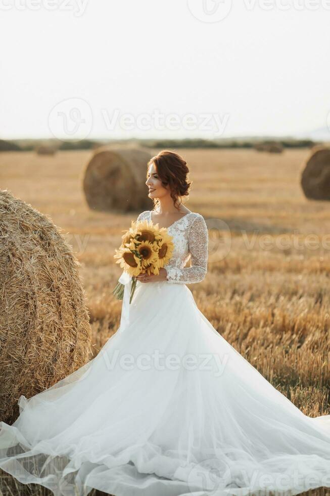 Wedding portrait. A red-haired bride in a white dress, holding a bouquet of sunflowers, on the background of a field. Beautiful curls. Sincere smile. Elegant dress. Yellow flowers photo