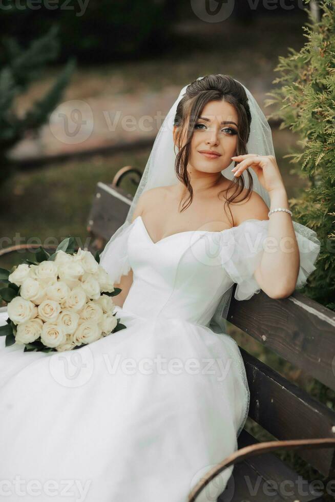 Portrait of the bride in nature. Brunette bride in a white long dress, holding a bouquet of white roses, sitting on a bench, posing, looking into the lens. on the background of the tree. Curly hair photo