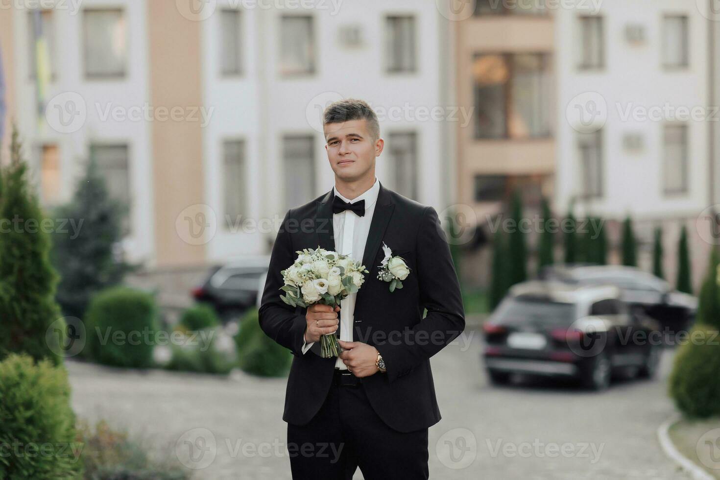 A man in a white shirt, a black bow tie and a black suit holds a bouquet and goes to a meeting, behind him are black cars, new buildings and trees. A stylish watch. Men's style. Fashion. Business photo