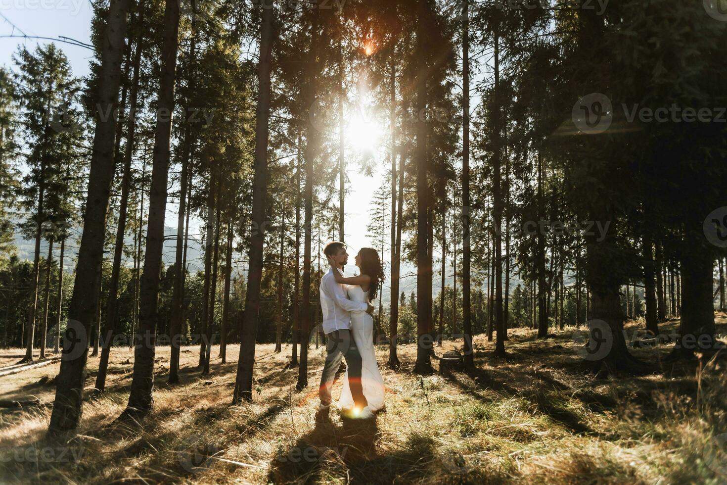 Stylish model couple in white clothes kissing at sunset in the forest. A girl hugs her husband, hair blowing in the wind. Wide angle photo