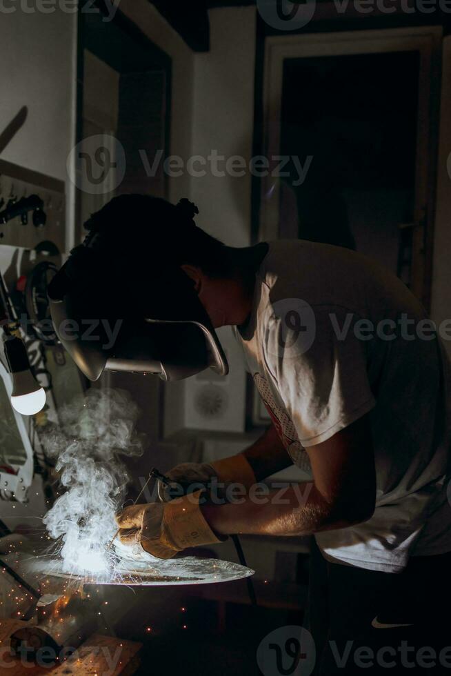 A welder is doing welding work in his garage while sparks fly around, he is wearing a safety helmet. Manual work with a welding device photo