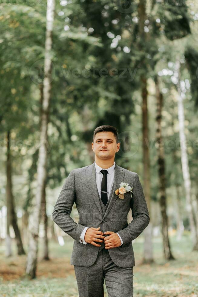 a stylish groom walks forward in the forest buttoning his jacket between tall trees. A fashionable groom, an adult man photo