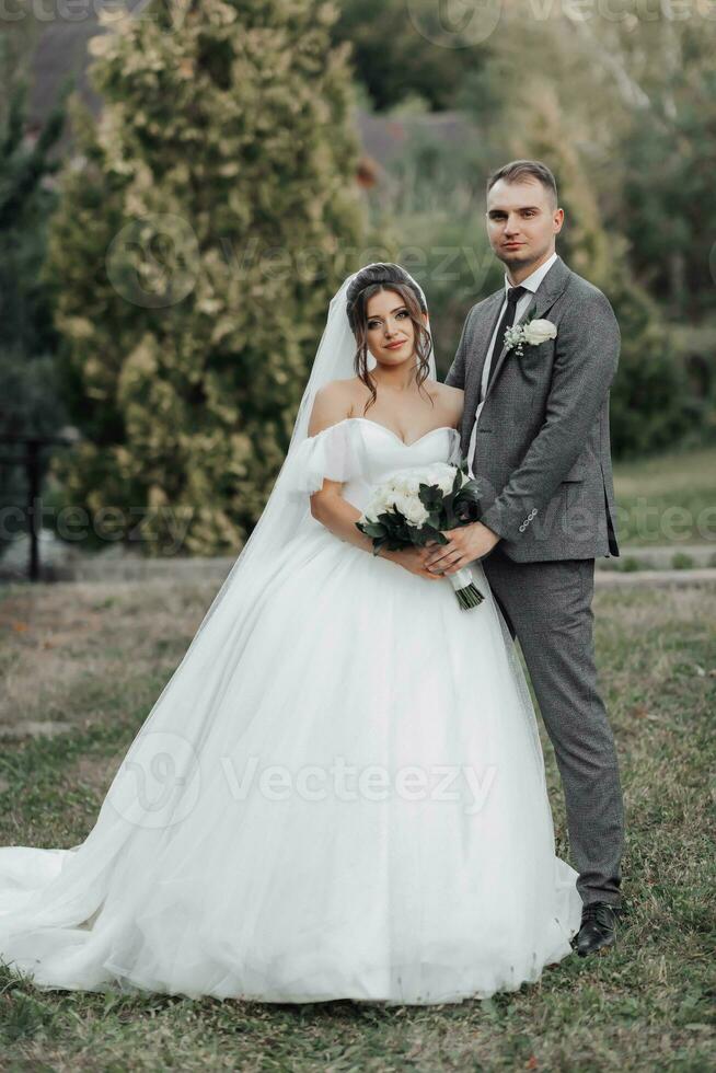 Portrait of the bride and groom in nature. Cropped photo. The bride and groom pose, hugging and smiling, against the background of conifers. The bride in a long dress with a bouquet of roses. photo