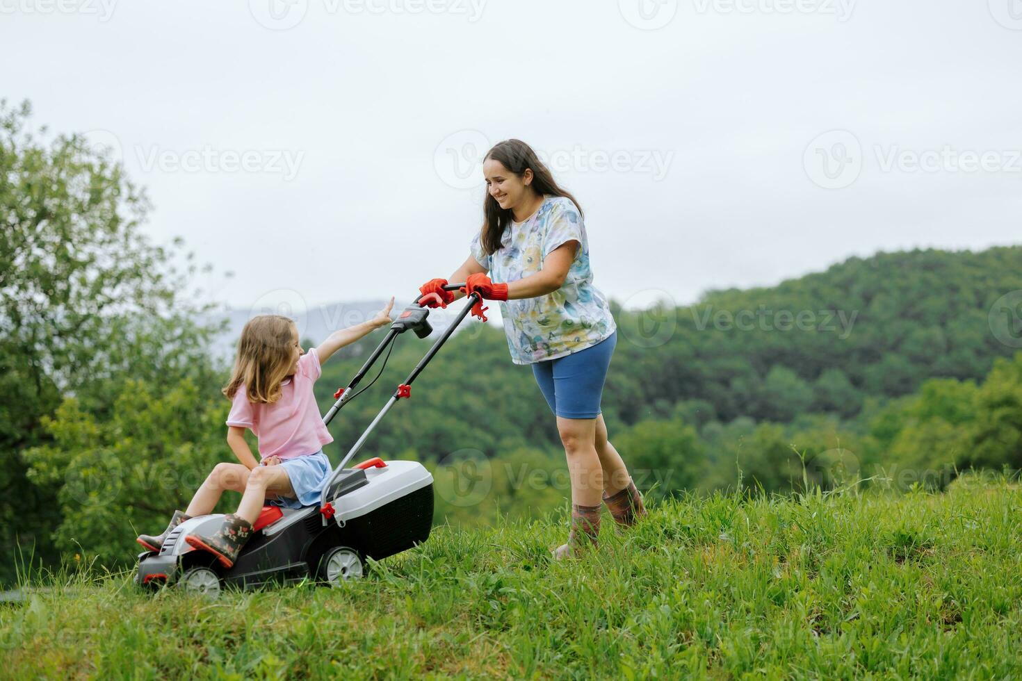 A woman in boots with her child in the form of a game mows the grass with a lawnmower in the garden against the background of mountains and fog, garden tools concept photo