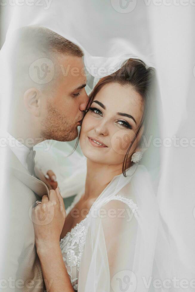 Wedding portrait of the bride and groom. Happy bride and groom gently hug each other under the veil, pose and kiss. Stylish groom. Beautiful young bride photo