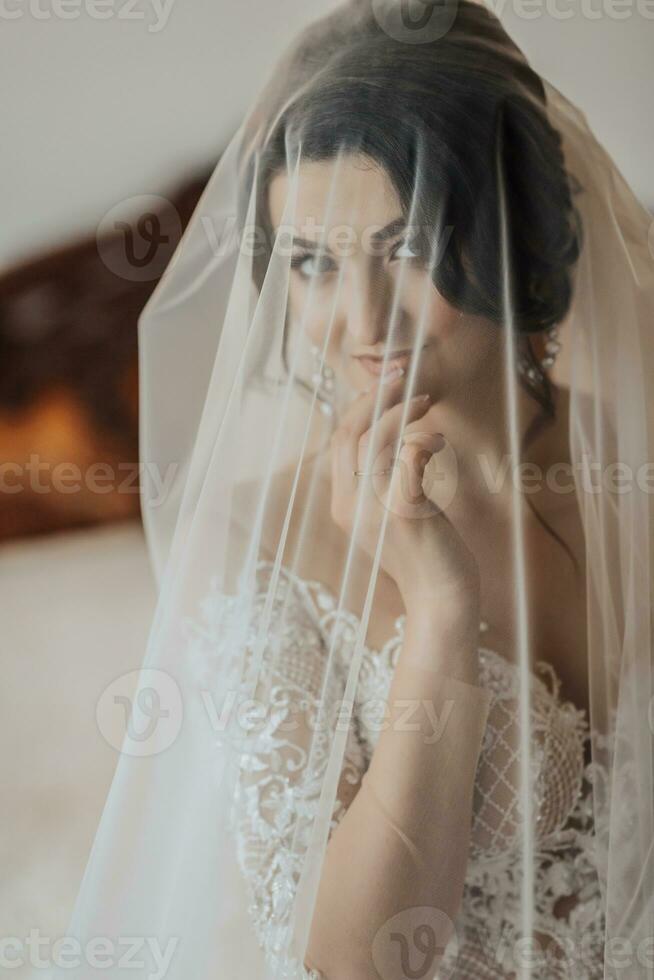 Portrait. A brunette bride in a white dress, wrapped in a veil, looks down and touches her face, poses. Gorgeous make-up and hair. Voluminous veil. Wedding photo. Beautiful bride photo