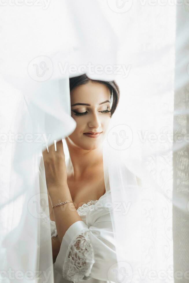 Portrait. A brunette bride is standing wrapped in a veil by the window and looking down. Gorgeous make-up and hair. Voluminous veil. Wedding photo. Beautiful bride photo