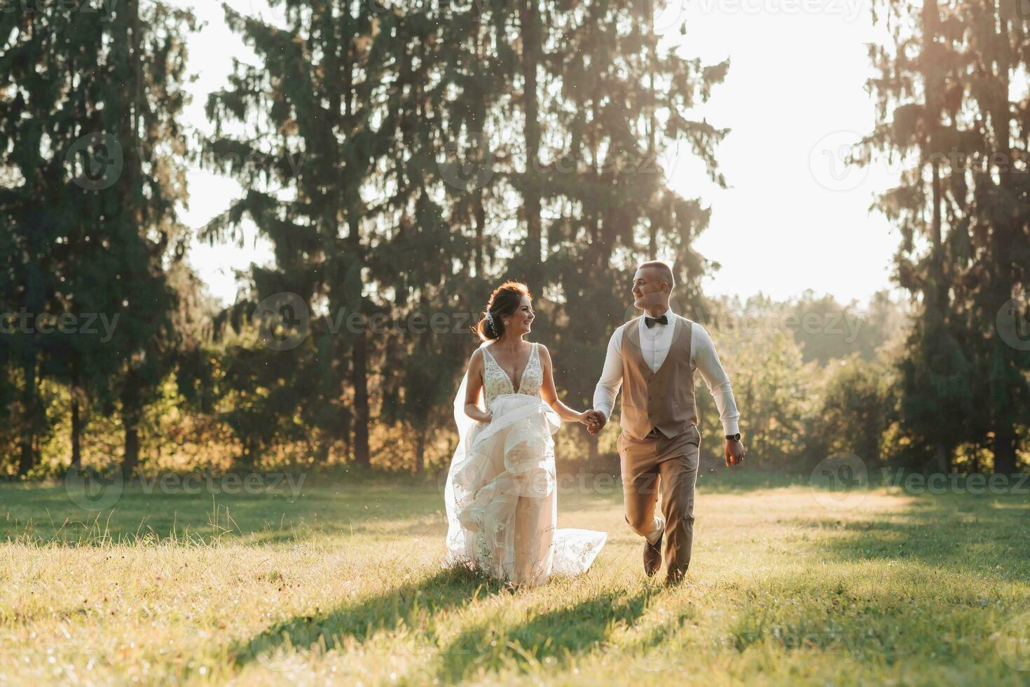 Happy newlyweds are running in the field, holding hands, looking at each other, behind them are big green Christmas trees. Beautiful light. Stylish groom. Pretty Girl photo