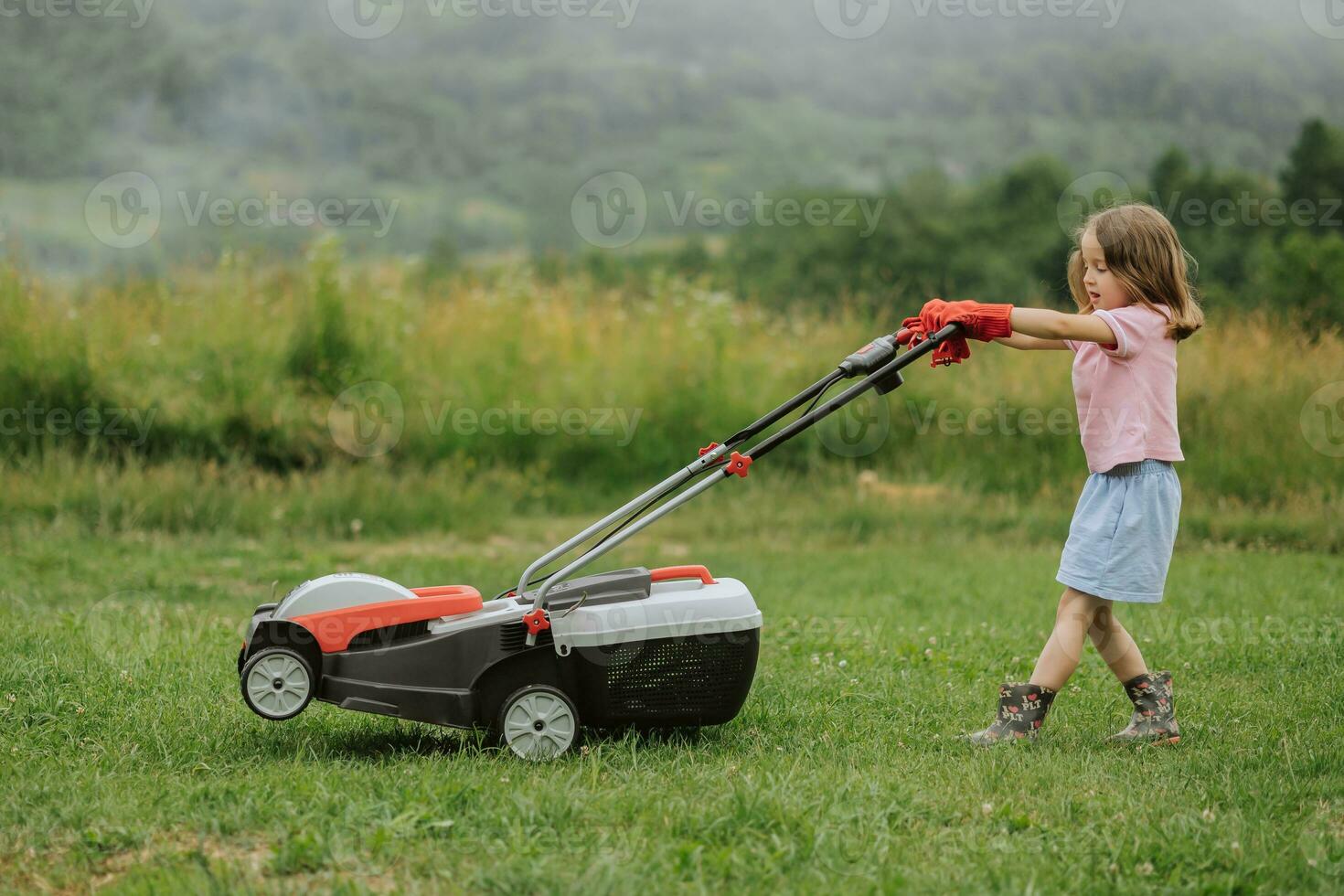 A child in boots in the form of a game mows grass with a lawnmower in the yard against the background of mountains and fog, the concept of garden tools photo