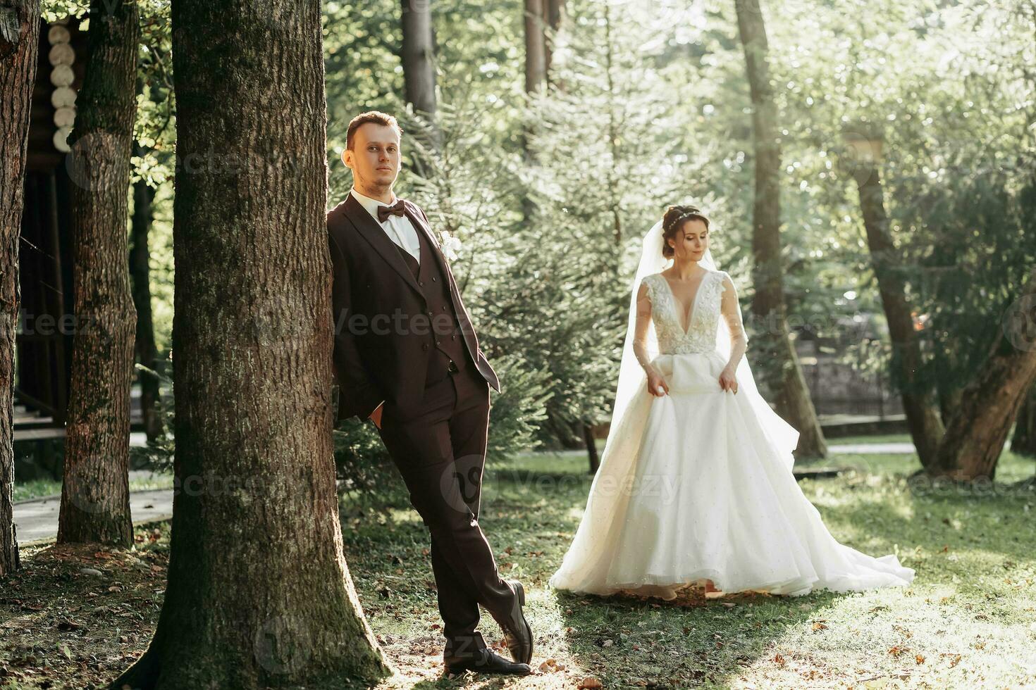 full-length portrait of a young bride and groom walking on a green golf course at sunset. Happy wedding couple, space for text. The groom leans against a tree, the bride is behind photo