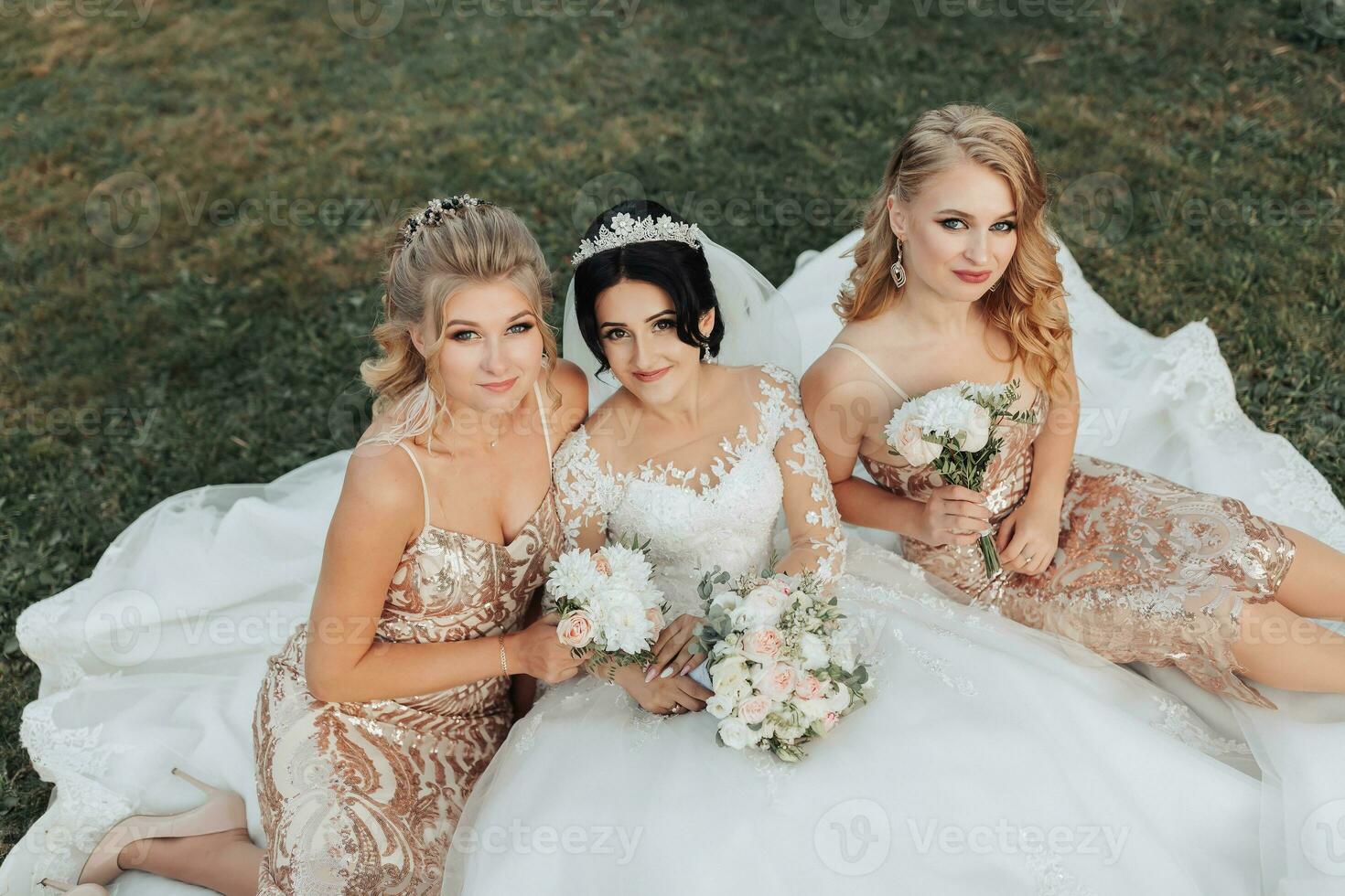 A brunette bride in a white elegant dress with a crown and her blonde friends in gold dresses pose with bouquets while sitting on the grass. Wedding portrait in nature, wedding photo in a light tone.