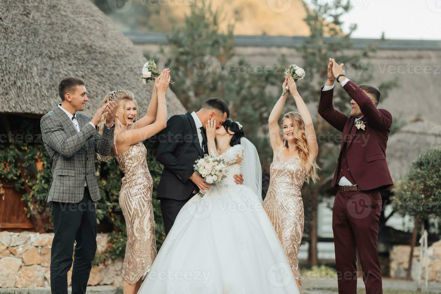 Front view of newlyweds standing and kissing, between friends, friends having fun. Girls in golden dresses hold bouquets, the bride in a voluminous dress, the groom and his friends in suits photo