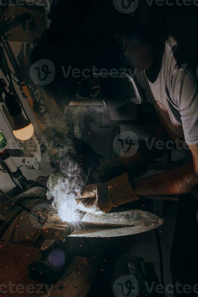 A welder is doing welding work in his garage while sparks fly around, he is wearing a safety helmet. Manual work with a welding device photo