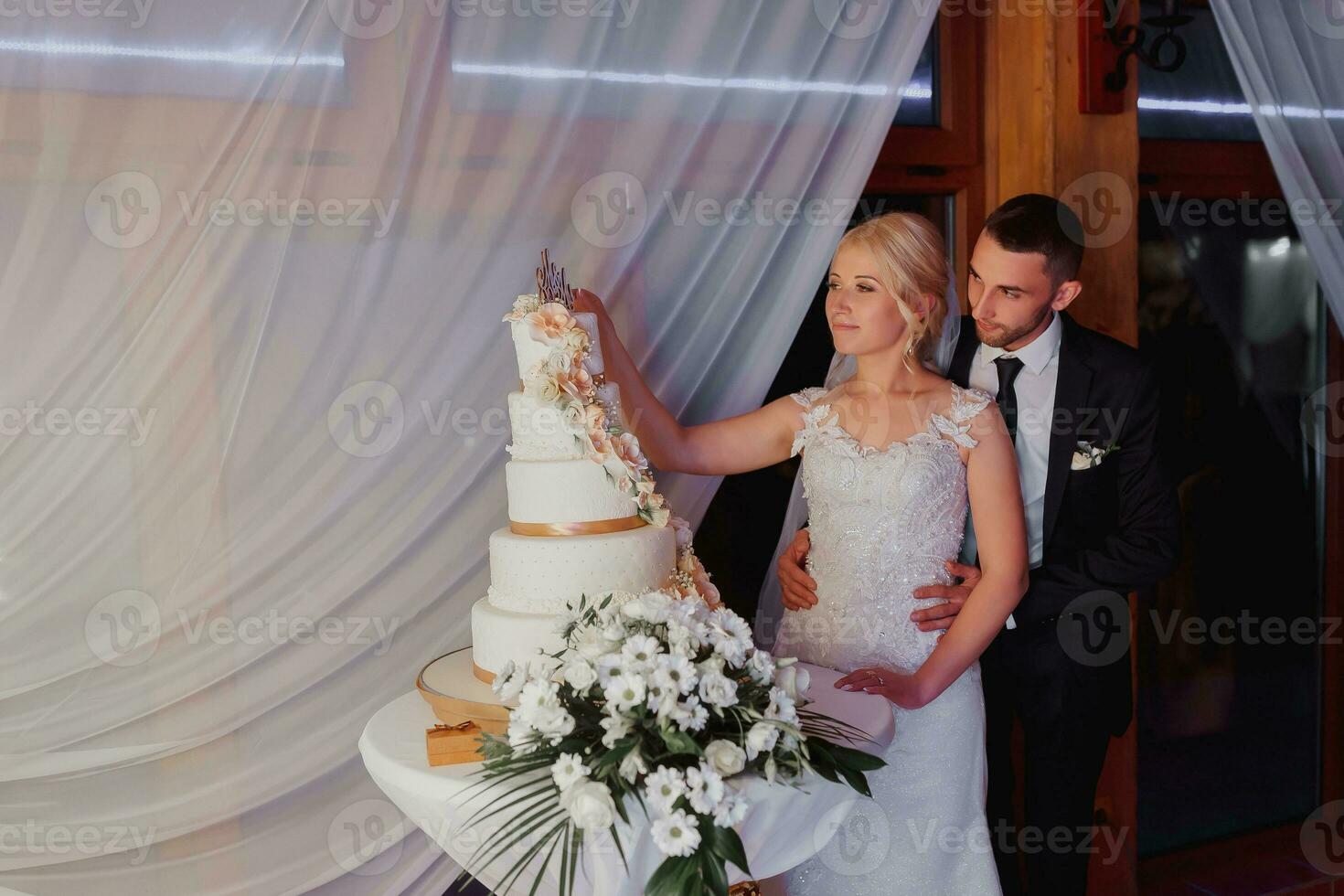 A white wedding cake, decorated with flowers and gold, stands on a glass stand. Fresh flowers for a wedding. sweets. The bride and groom near the cake photo