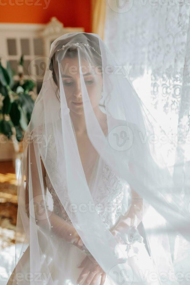 A brunette bride in a white dress holds her wedding bouquet, poses. French manicure. Open shoulders. Beautiful hands. Long veil. Morning of the bride. Details photo