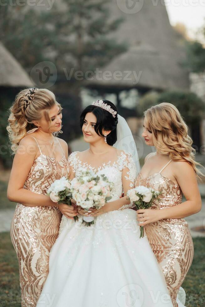 un morena novia en un blanco elegante vestir con un corona y su rubia amigos en oro vestidos actitud con ramos de flores Boda retrato en naturaleza, Boda foto en un ligero tono.