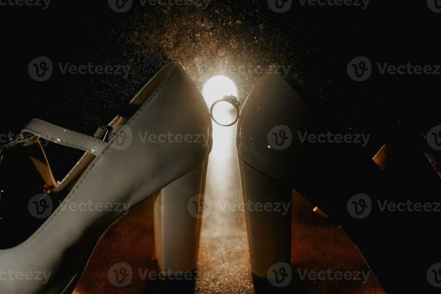 Photo of details at the wedding. An engagement ring with a diamond stands between white shoes, leather sandals with decor, on the floor in the backlight.