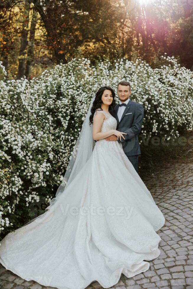Happy young couple, bride with long curly hair in white dress with long train near castle in beautiful flowers. Beautiful girl in the park. Beautiful sunlight. Wedding photo shoot