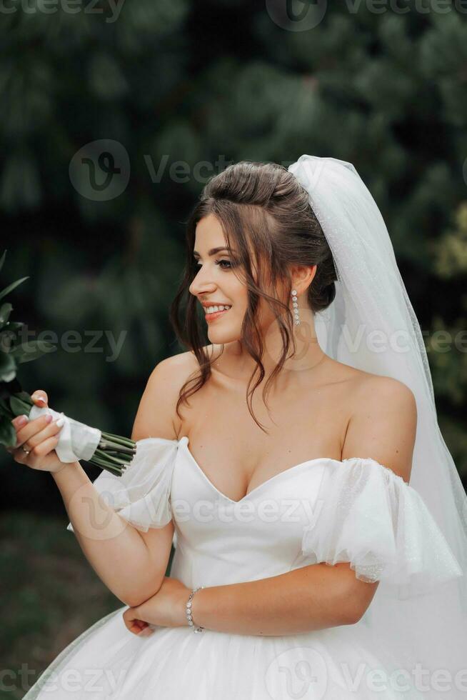 Brunette Bride in a voluminous white dress with open shoulders and a long veil, smiling, posing with a bouquet of white roses. Portrait of the bride. Beautiful makeup and hair. photo