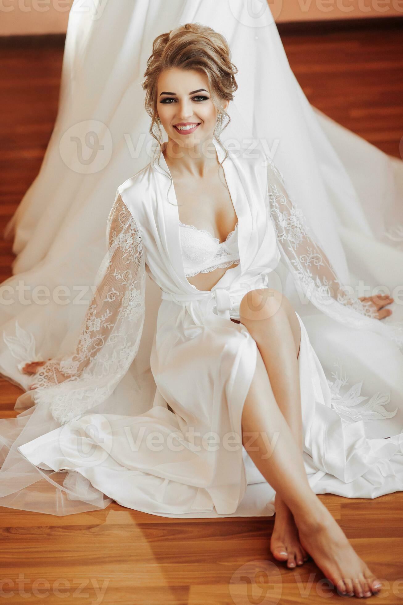 A blonde bride in a long lace petticoat and white shoes sits in her room  near her wedding dress, poses. Beautiful hair and makeup, open bust.  Wedding portrait. Open legs 36424038 Stock