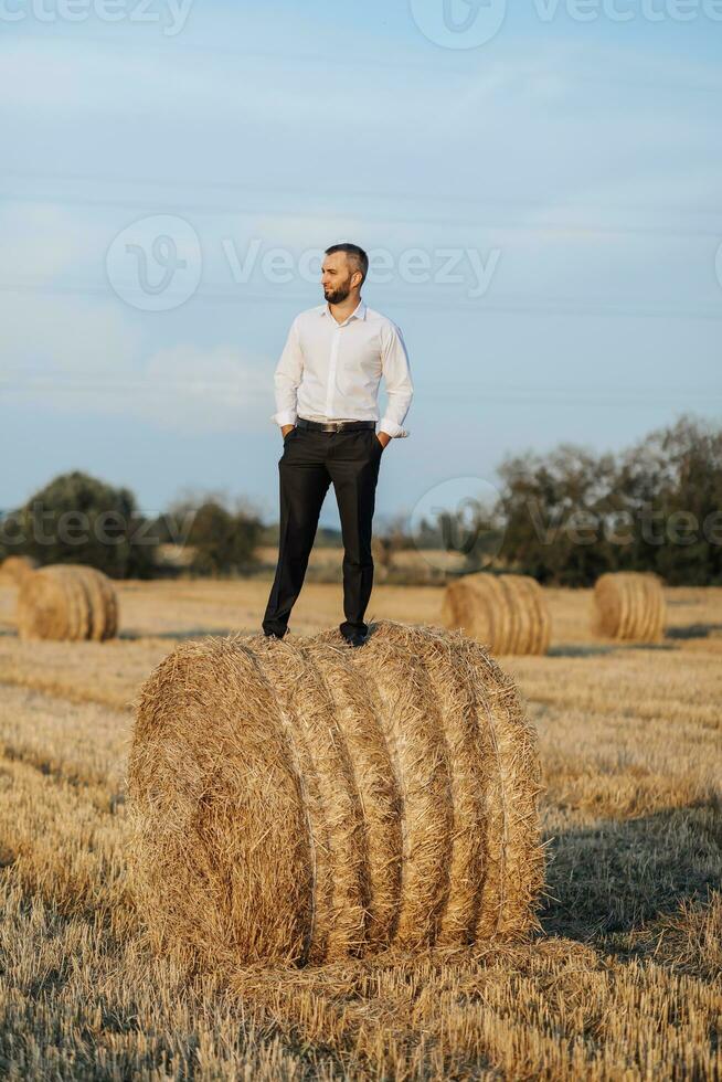 Boda retrato foto. un elegante novio en un blanco camisa poses en un heno bala con su manos en su bolsillos barbado hombre. estilo. foto